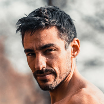 Man with short dark hair and beard looking directly at the camera with a blurred natural background