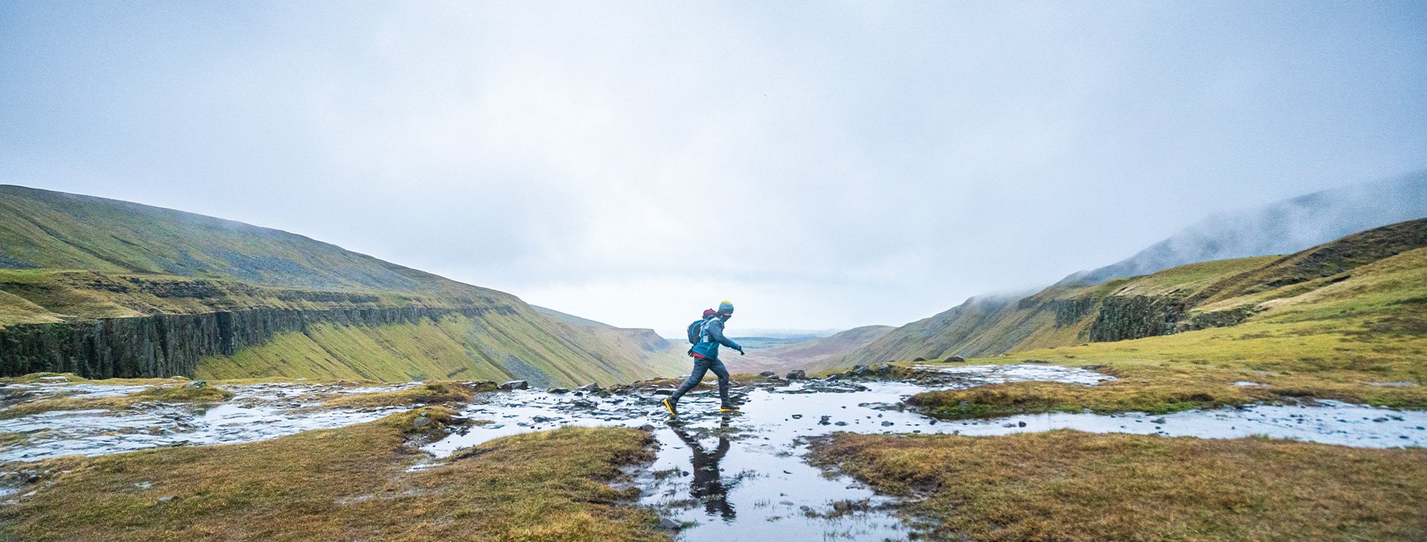 Person hiking through a mountainous landscape with green grass and a clear sky.