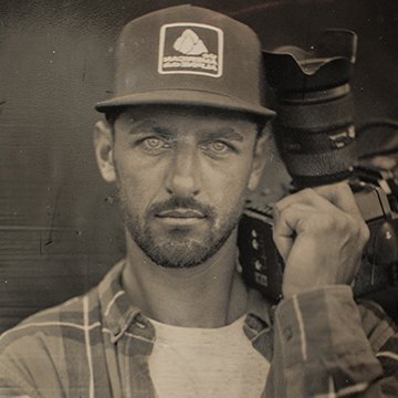 Person wearing a cap with a logo, holding a camera, in a black and white photo.