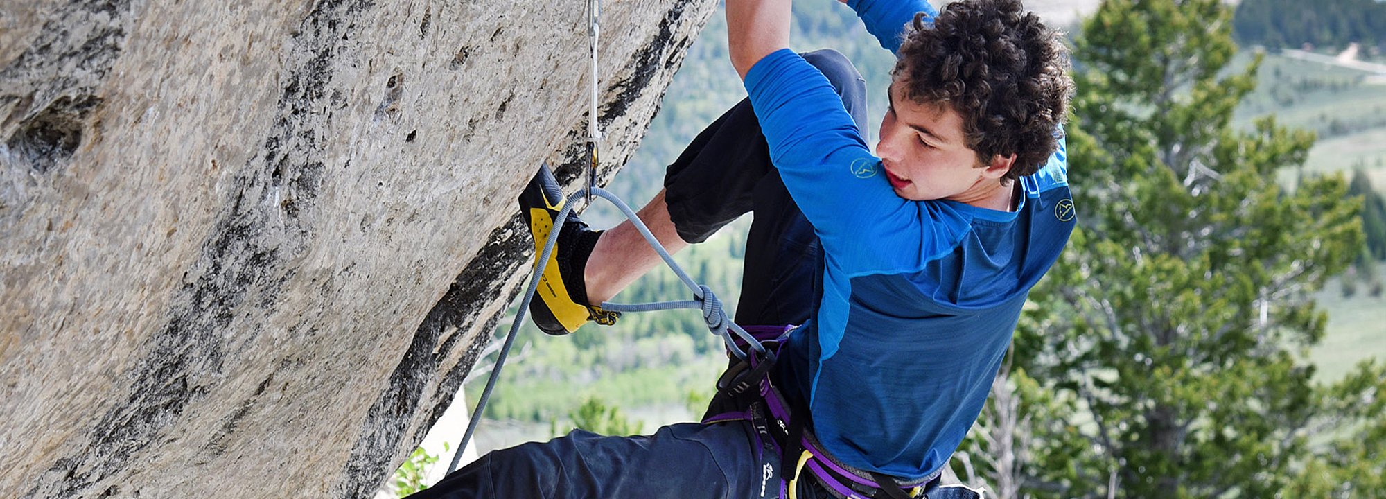 A young man with curly hair, wearing a blue shirt and climbing gear, ascends a steep outdoor rock face. Green trees and hills are visible in the blurred background.