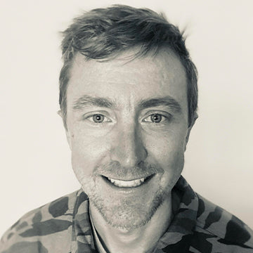 Black and white portrait of a man with tattoos on his arm, wearing a patterned shirt.