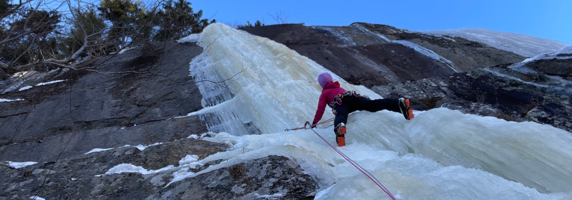 A person in winter climbing gear ascends a steep, frozen waterfall on a rocky mountainside, using ice axes and crampons under a clear blue sky.