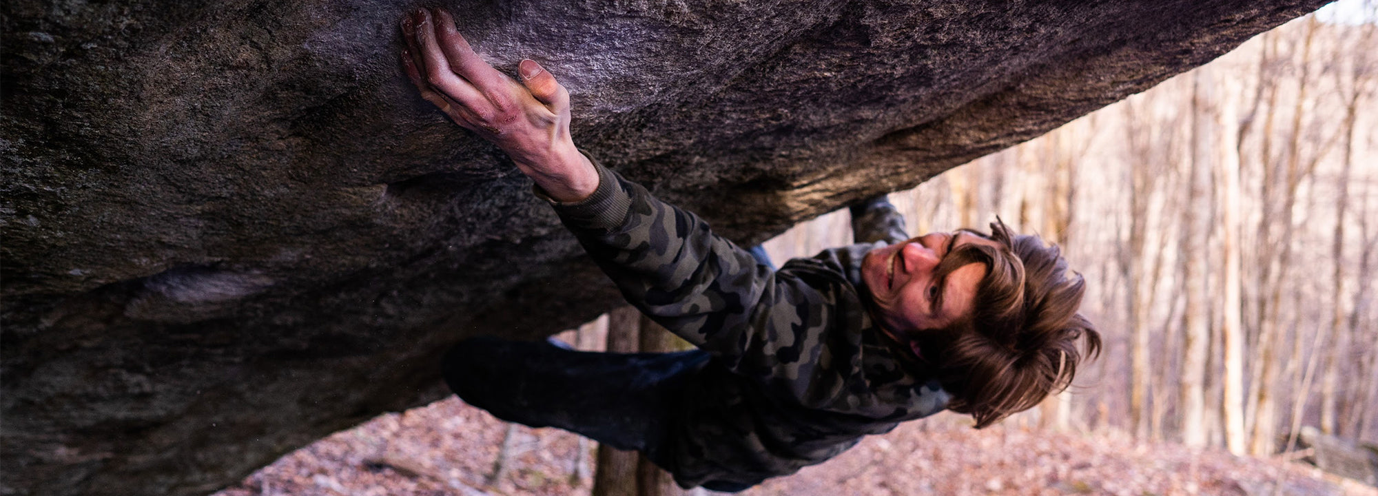 A person with brown hair and a camouflage jacket climbs a steep, overhanging rock outdoors, gripping a hold with one hand. Leafless trees fill the background, suggesting autumn or winter.