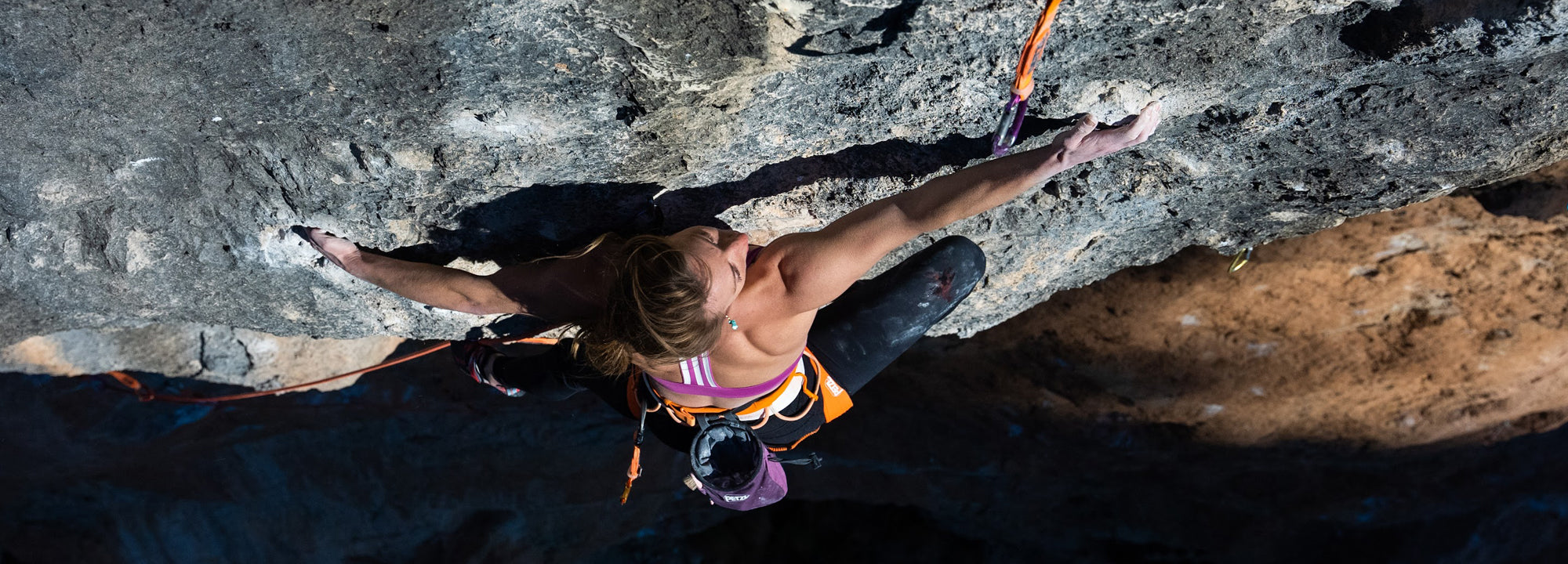 A woman wearing a harness and athletic clothing climbs a steep, rocky cliff face, gripping small holds with her hands and feet while secured by ropes. Sunlight highlights the rock’s rough texture.