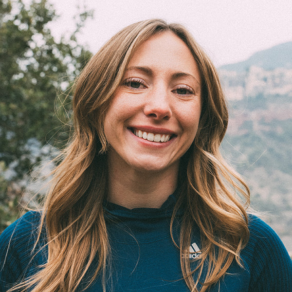 Woman with long blonde hair wearing a blue Adidas shirt outdoors with trees and mountains in the background.