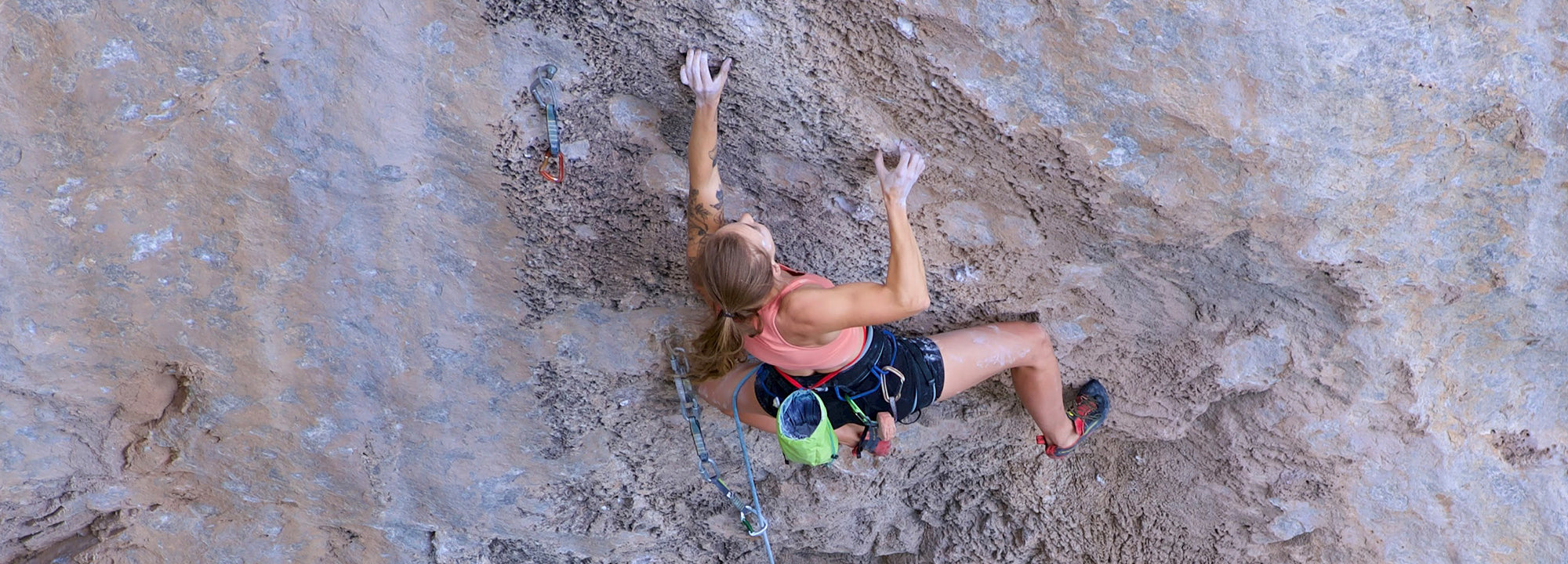 A climber wearing a pink tank top and black shorts ascends a rocky cliff face, using climbing gear and chalk. The image is taken from above, showing her focused expression and athletic posture.