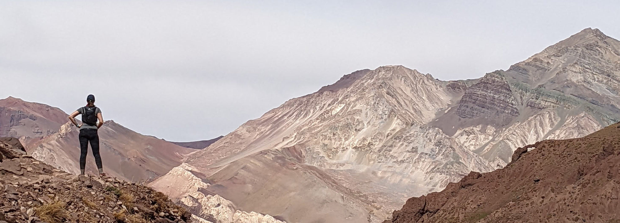 A person wearing a backpack stands on a rocky hill, looking out at a vast, rugged mountain range under a cloudy sky.