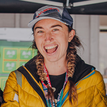 Person wearing a yellow jacket and cap with braided hair, smiling outdoors.