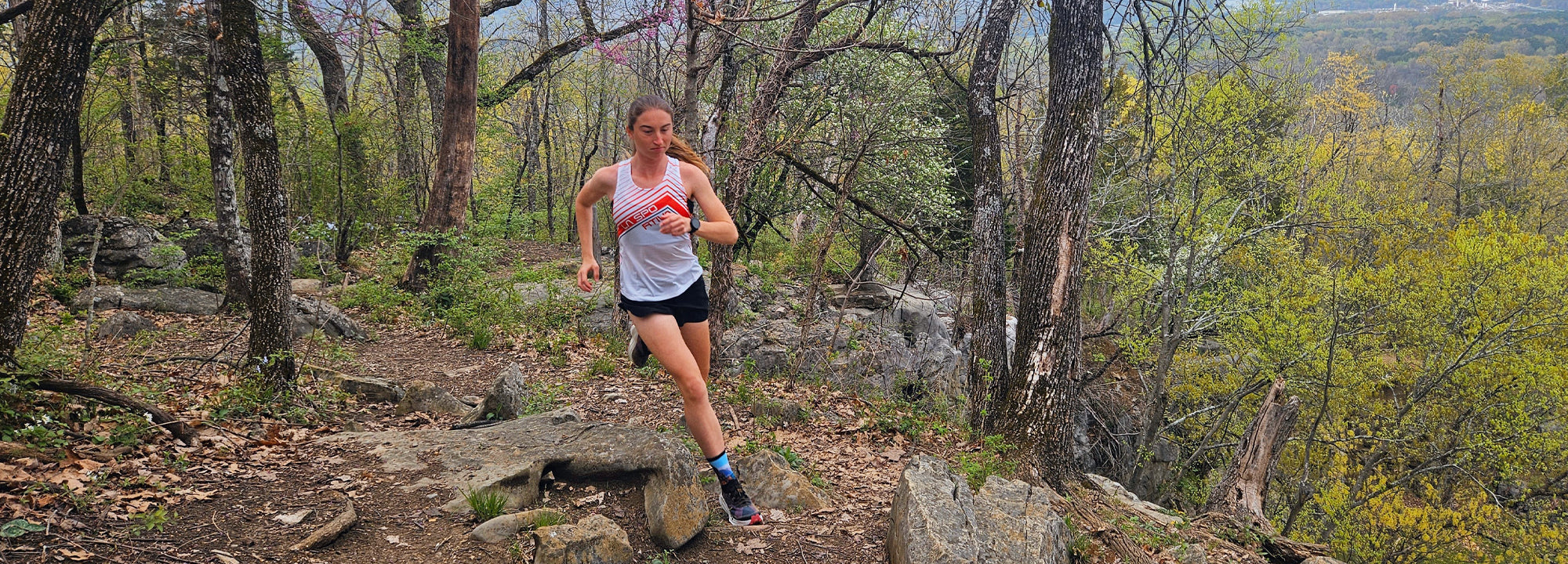 A woman in athletic gear runs on a rocky, wooded trail surrounded by trees and greenery, with a distant view of hills and cloudy sky in the background.