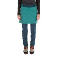 A person models the La Sportiva Warm Up Primaloft Skirt Women's in teal over blue pants with mountaineering boots, standing against a white background—their upper body partly out of frame, hands relaxed, creating an après ski vibe.