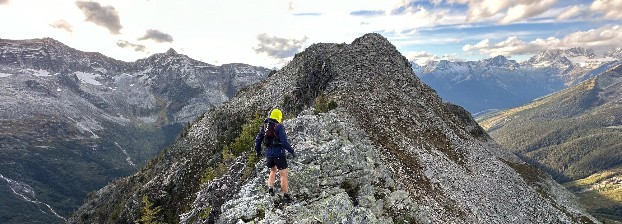 Person on a mountain peak with a backpack and helmet, surrounded by snow-capped mountains.