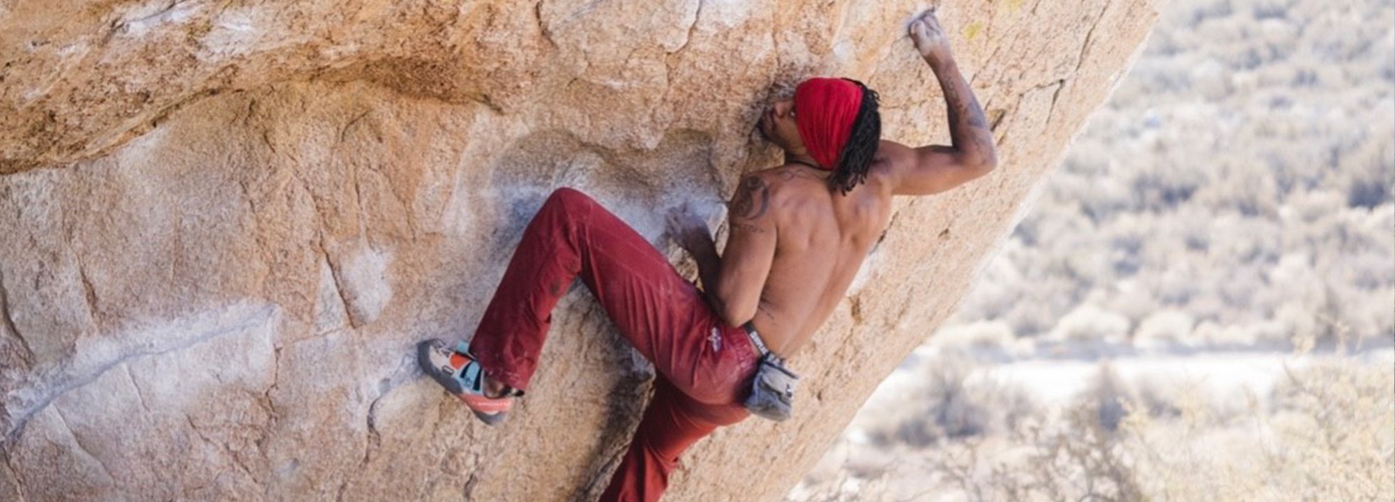 A person wearing red pants, a red bandana, and climbing shoes is bouldering shirtless on a large, steep rock face outdoors. The background is slightly blurred, showing dry, rocky terrain.