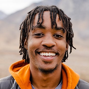 Man with dreadlocks wearing an orange hoodie against a mountainous background