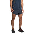 A person stands facing forward with arms relaxed by their sides, wearing a navy blue T-shirt, La Sportiva Auster Shorts Men's, patterned socks, and black athletic shoes. Their head is not visible in the image.
