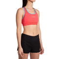 Une femme présente le La Sportiva Hover Top Women's, une brassière de sport rouge avec des bordures bleues, associée à un short d'athlétisme noir, en se tenant légèrement de biais sur un fond blanc.