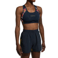 Une personne portant la brassière de sport La Sportiva Hover Top Women avec des bretelles géométriques colorées et un short marine assorti avec des panneaux latéraux jaunes, debout sur un fond blanc.