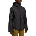 Une femme portant la Discover Shell Jacket Women's (2025) en gris foncé, avec une capuche réglable et une fermeture à glissière sur le devant, associée à un pantalon jaune moutarde, se tient sur un fond blanc uni.