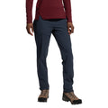 Une personne porte le West Crest Pants Women's (2025) de La Sportiva, qui est un pantalon de randonnée bleu foncé avec des poches à fermeture éclair. Elle porte également une chemise à manches longues bordeaux et des chaussures de randonnée marron. Elle se tient sur un fond blanc, sa tête n'est pas visible.
