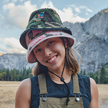 Person wearing a colorful bucket hat and overalls with a mountainous background