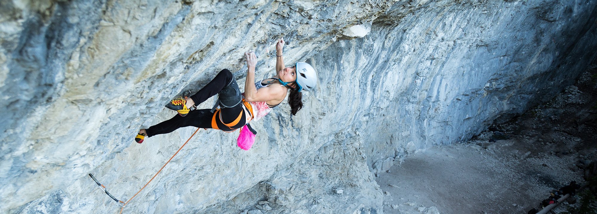 A woman wearing a helmet and harness climbs a steep, rocky cliff using ropes. She grips the rock with one hand and extends her legs, appearing focused and determined, with climbing gear attached to her harness.