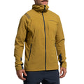 A man stands with hands at his sides, wearing the La Sportiva Aequilibrium Speed Jacket Men's (2025)—a mustard yellow, lightweight hooded jacket with a front zipper, dark blue accents, and branded chest detailing.