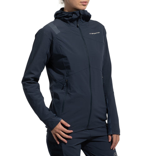Une femme porte la veste La Sportiva Aequilibrium Speed Jacket (2025) en marine foncé, avec une capuche, une fermeture éclair, des poches, un petit logo sur la poitrine et un design subtil sur les épaules. La veste respirante est associée à un pantalon assorti sur fond blanc.