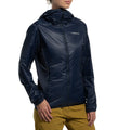 A person wearing the La Sportiva Aequilibrium Lite Insulation Jacket Women's (2025), a dark blue, breathable, warm jacket with a front zipper, and light brown pants stands against a white background. Only the upper body and part of the face are visible.