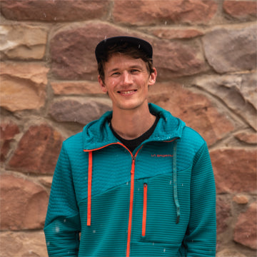 Person wearing a teal jacket with orange accents standing in front of a stone wall.