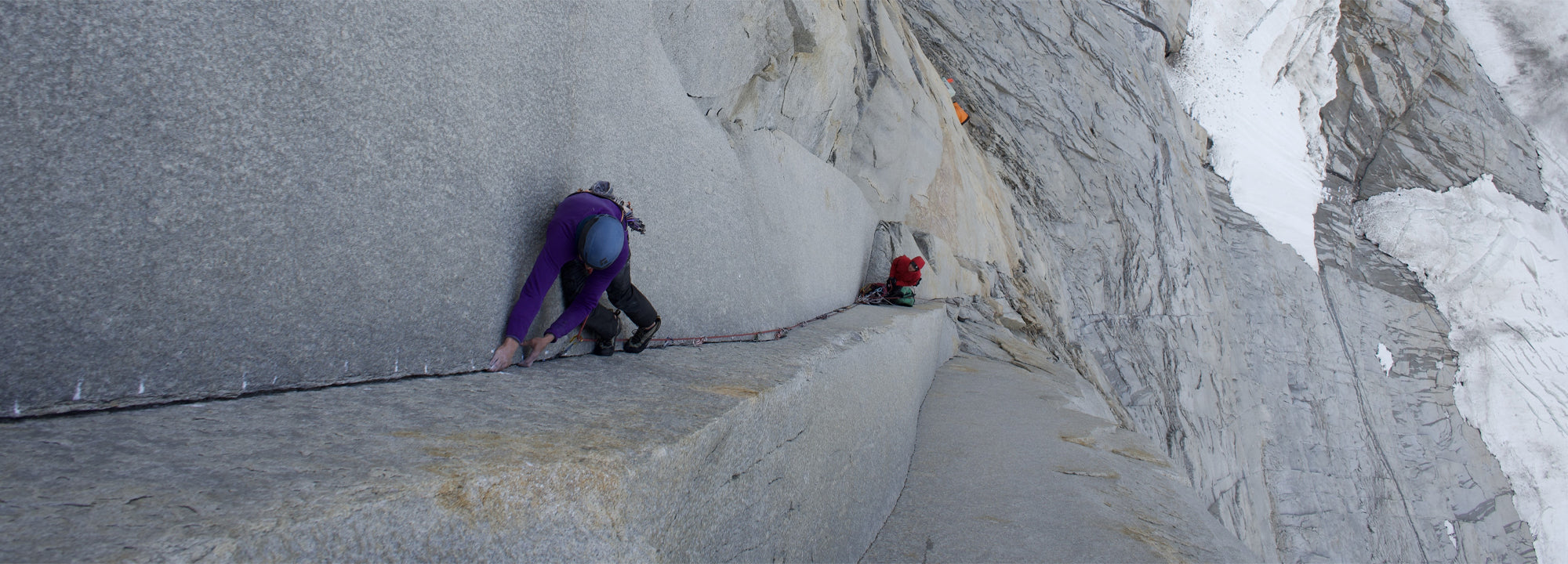 Two climbers ascend a steep, narrow granite ledge on a mountain face. One climber in a purple jacket crawls along the rock, while the other in red follows, secured by ropes. Snow-covered peaks are visible in the background.