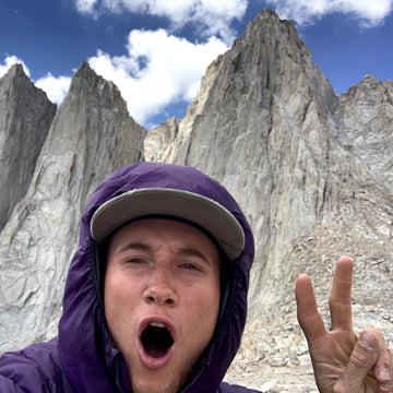 Person in a purple jacket and cap posing in front of mountain peaks with a blue sky.