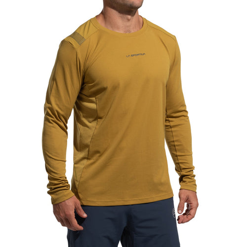 Une personne porte un haut La Sportiva Beyond Long Sleeve Men's (2025) jaune moutarde avec un tissu léger Polartec Delta Cooling Fabric et un pantalon bleu marine, debout, les bras détendus sur un fond blanc. La tête est partiellement hors du cadre.