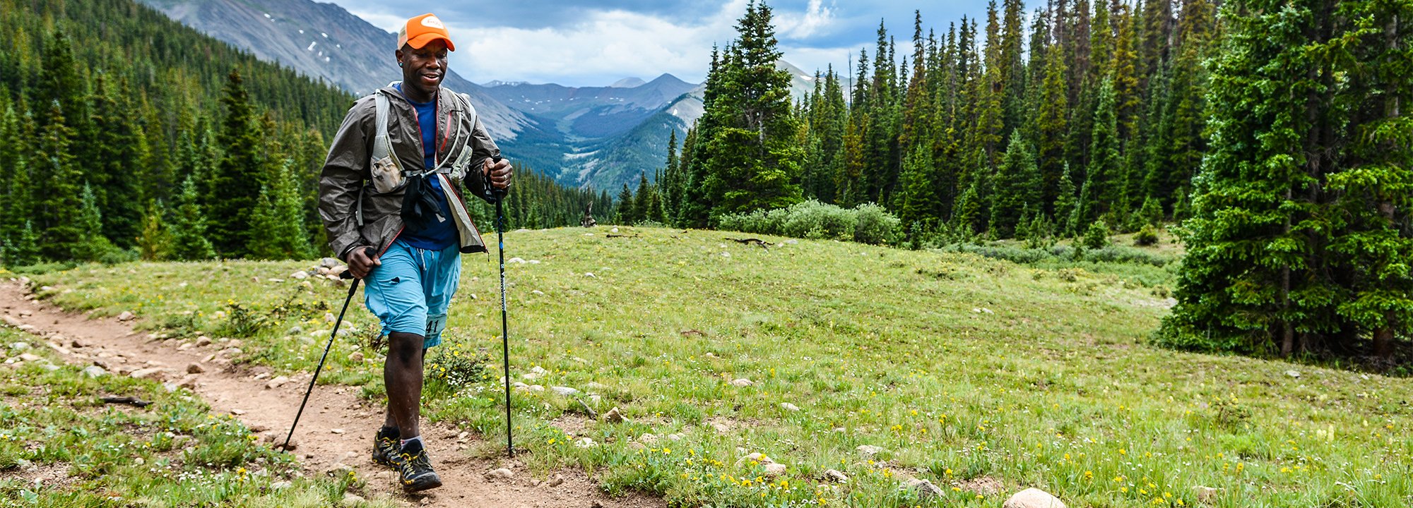 A person wearing an orange cap and blue shorts hikes on a mountain trail with trekking poles, surrounded by green trees and distant mountain peaks under a cloudy sky.