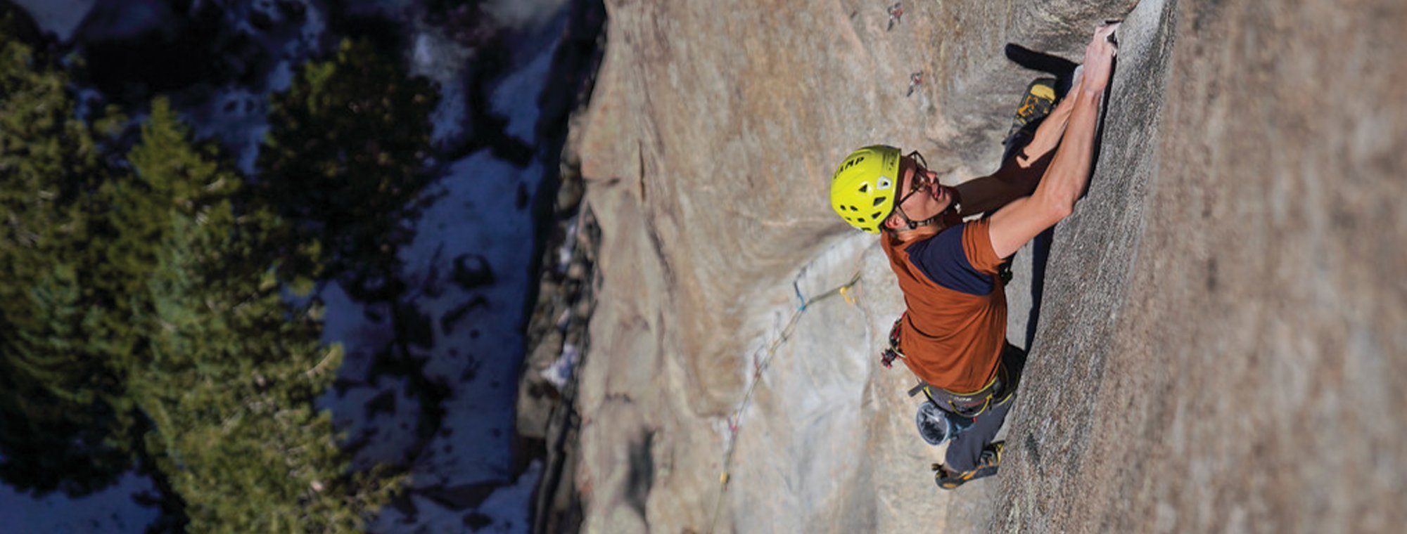 A rock climber wearing a yellow helmet and orange shirt climbs a steep rock face, gripping a crack with one hand. Trees and shadows are visible far below.