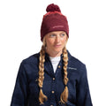 A woman with blonde braided hair wears the La Sportiva Knitty Beanie (2025), a maroon synthetic knit hat featuring a pom-pom, along with a navy blue plaid shirt. She looks slightly to her right and smiles softly, ready to take on the cold weather in style.