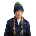 A woman with blonde braided hair is wearing the La Sportiva Terry Beanie Women's in navy blue with green accents and a pom-pom, paired with a dark blue plaid button-up shirt. She looks to the side and smiles softly.