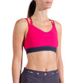 A woman models the La Sportiva Boulder Bra Women's in bright pink with double shoulder straps and dark gray high-waisted pants, standing against a white background.