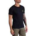Un homme portant le t-shirt La Sportiva Climbing on the Moon en noir, avec un petit logo jaune sur la poitrine et un pantalon vert olive. Il se tient debout sur un fond blanc, un bras tatoué visible, regardant légèrement vers le haut.