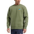 A person wearing the La Sportiva Moon Climb Sweater—a recycled climber’s sweater in olive green featuring a small colorful patch on the left chest—paired with navy blue pants, stands against a plain white background.