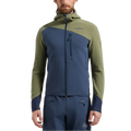 Un homme se tient debout, le regard tourné vers l'avant, les mains détendues le long du corps. Il porte le sweat à capuche thermique La Sportiva Bristen pour homme, une veste technique d'alpinisme conçue pour les climats froids, qui se distingue par ses manches et ses épaules vert olive contrastant avec le devant et la capuche bleus.