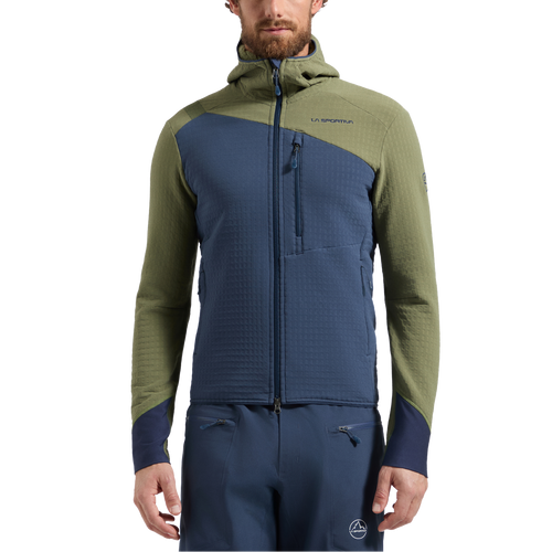A man stands facing forward with his hands relaxed by his sides, wearing the La Sportiva Bristen Thermal Hoody Men’s—a technical mountaineering jacket designed for cold climates, distinguished by its olive green sleeves and shoulders contrasted with a blue front and hood.