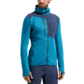 Un homme se tient légèrement de profil devant un fond blanc uni. Il porte le sweat à capuche thermique La Sportiva Lucendro pour homme, de couleur bleue et bleu marine — doté de passe-pouces, idéal comme couche thermique pour les alpinistes — ainsi qu'un pantalon bleu marine assorti.