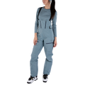 A woman wearing the La Sportiva Sender GTX Performance Bib Pants for women, styled with a blue-gray long-sleeve shirt, black-strap details, white sneakers with black accents, and a matching headband, stands against a plain white background.