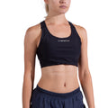 Une femme vêtue d'un soutien-gorge La Sportiva Pulse High Support Bra noir à bretelles croisées et d'un short d'athlétisme bleu marine se tient de face sur un fond blanc uni, son visage étant partiellement hors champ.