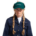 A woman with long blonde braids wears a green La Sportiva Forest Earflap Cap with sherpa lining, a navy plaid shirt, and a white undershirt, standing against a white background.