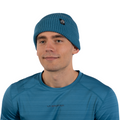 A man wearing a blue Boulder Beanie by La Sportiva and a blue La Sportiva shirt looks slightly to the side, smiling softly. The background is plain white.