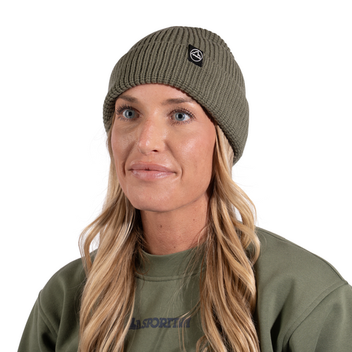 Une femme aux longs cheveux blonds porte le bonnet « Boulder Beanie » de La Sportiva, de couleur vert olive, ainsi qu'un sweat-shirt assorti. Elle regarde légèrement sur le côté et sourit doucement, avec un fond blanc derrière elle.