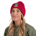 A woman with long blonde hair wears the La Sportiva Climbing on the Moon Beanie—a red knit hat featuring a colorful patch—paired with a green sweatshirt, radiating timeless style and warmth as she gazes off to the side against a white background.