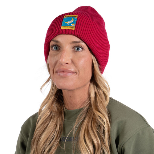 Une femme aux longs cheveux blonds porte le bonnet « Climbing on the Moon » de La Sportiva — un bonnet en tricot rouge orné d’un écusson coloré —, assorti à un sweat-shirt vert. Elle dégage une élégance intemporelle et une grande chaleur tandis qu’elle regarde au loin, sur un fond blanc.