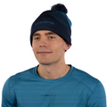 Un homme portant le bonnet Knitty Beanie de La Sportiva — un bonnet en tricot bleu orné d'un pompon — ainsi qu'un t-shirt de sport bleu assorti de la même marque est tourné vers l'avant et affiche un léger sourire sur fond blanc. Le style élégant de sa tenue en fait un choix idéal pour les journées froides.