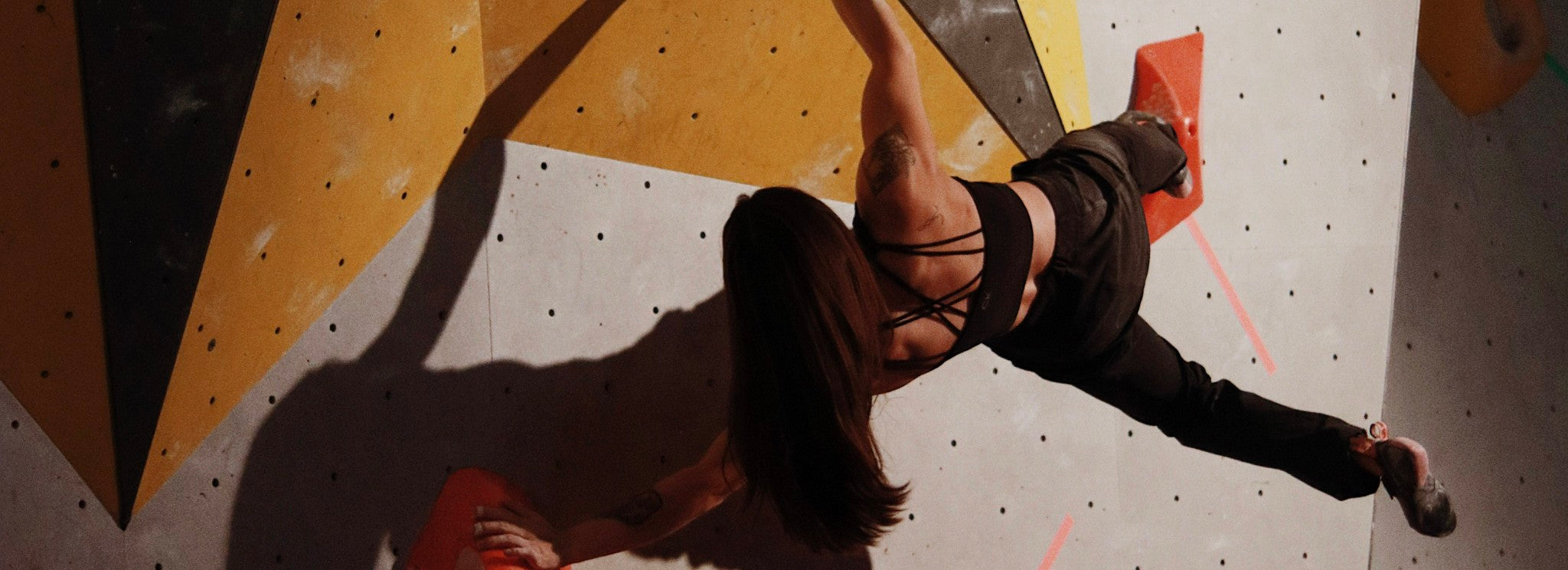 A woman with long hair climbs an indoor bouldering wall, reaching across large red holds. She wears a strappy black top and dark pants, with one leg extended and her back facing the camera.